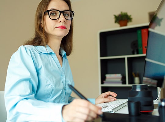 Frau mit Brille sitzt am Schreibtisch und zeichnet etwas auf einem Grafiktablett